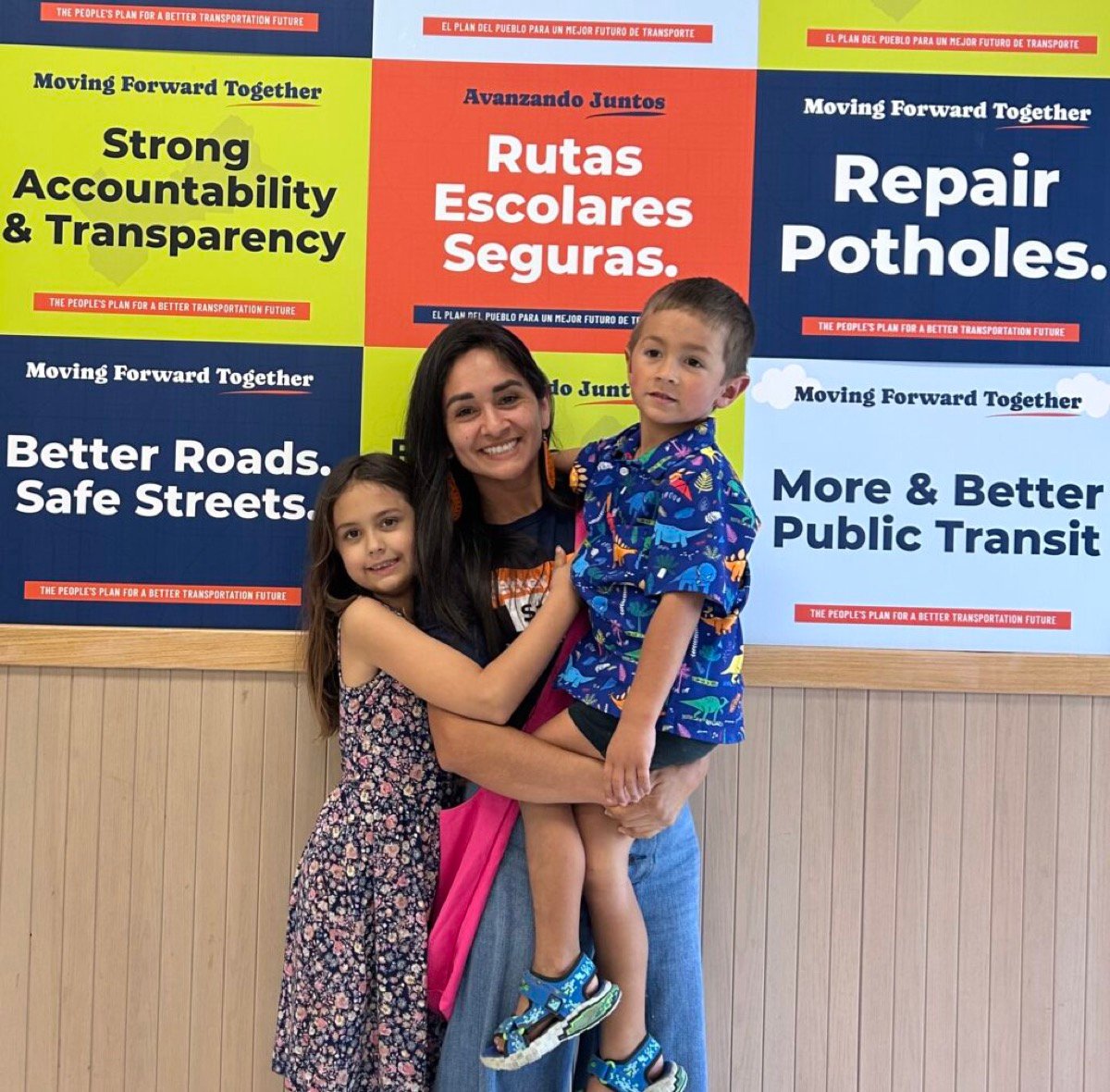 Ariana with her two children in front of bilingual community transit posters reading Better Roads, Safe Streets and Rutas Escolares Seguras