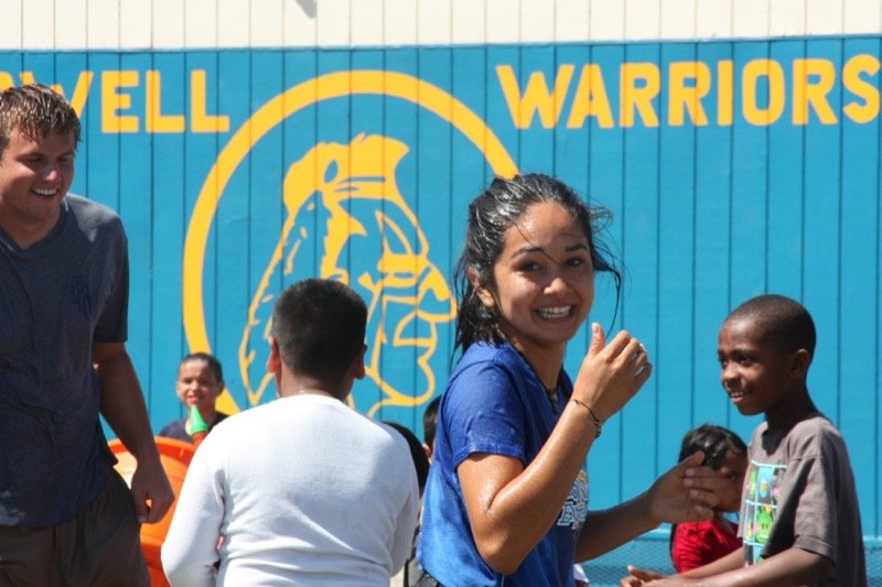 Ariana laughing with neighborhood children at a summer water event in front of the Lowell Warriors mural